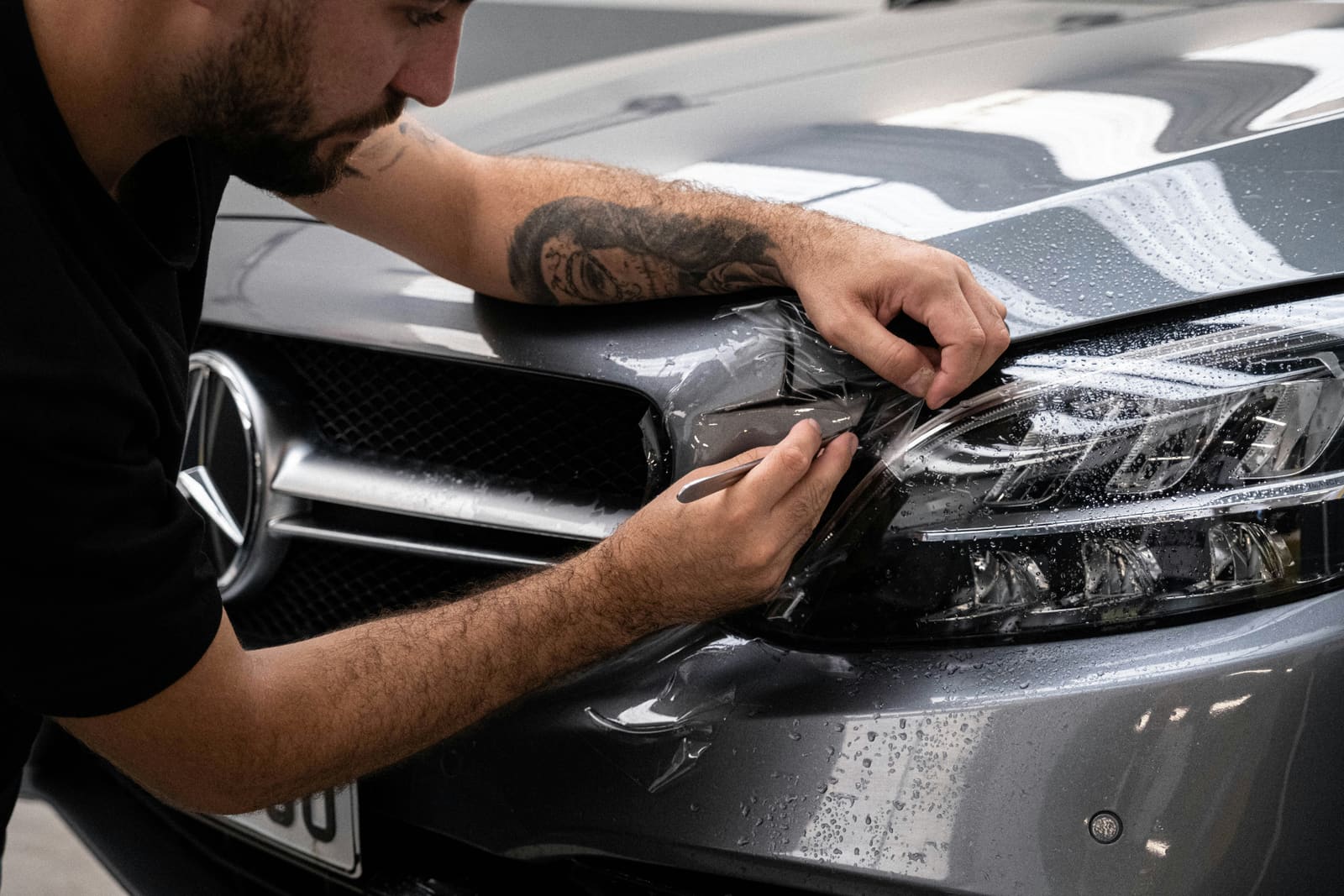 A technician's gloved hands squeegeeing a sheet of protective film flat around a car's headlight and front bumper edge.