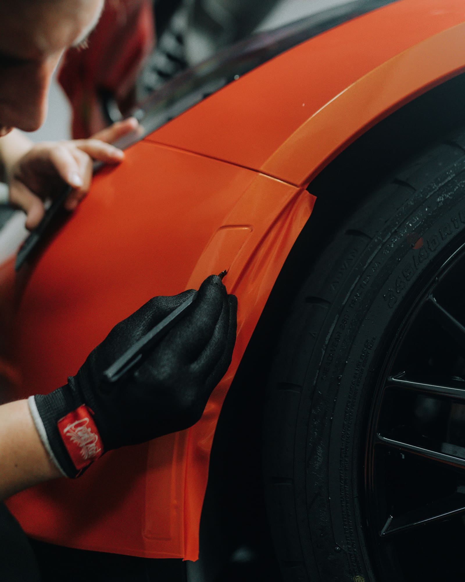 A detailer working the front fender and wheel of an orange car in a workshop.