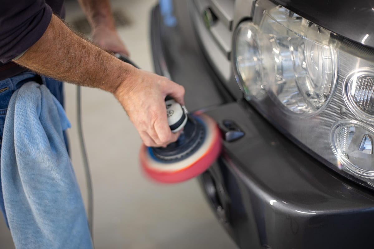A technician working a rotary polisher with a red foam pad along a car's front bumper, directly beside the clear headlight lens.
