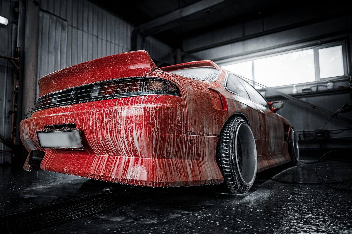 A car covered in soap suds during a wash in a detailing bay, rear bumper and taillight visible under running water.