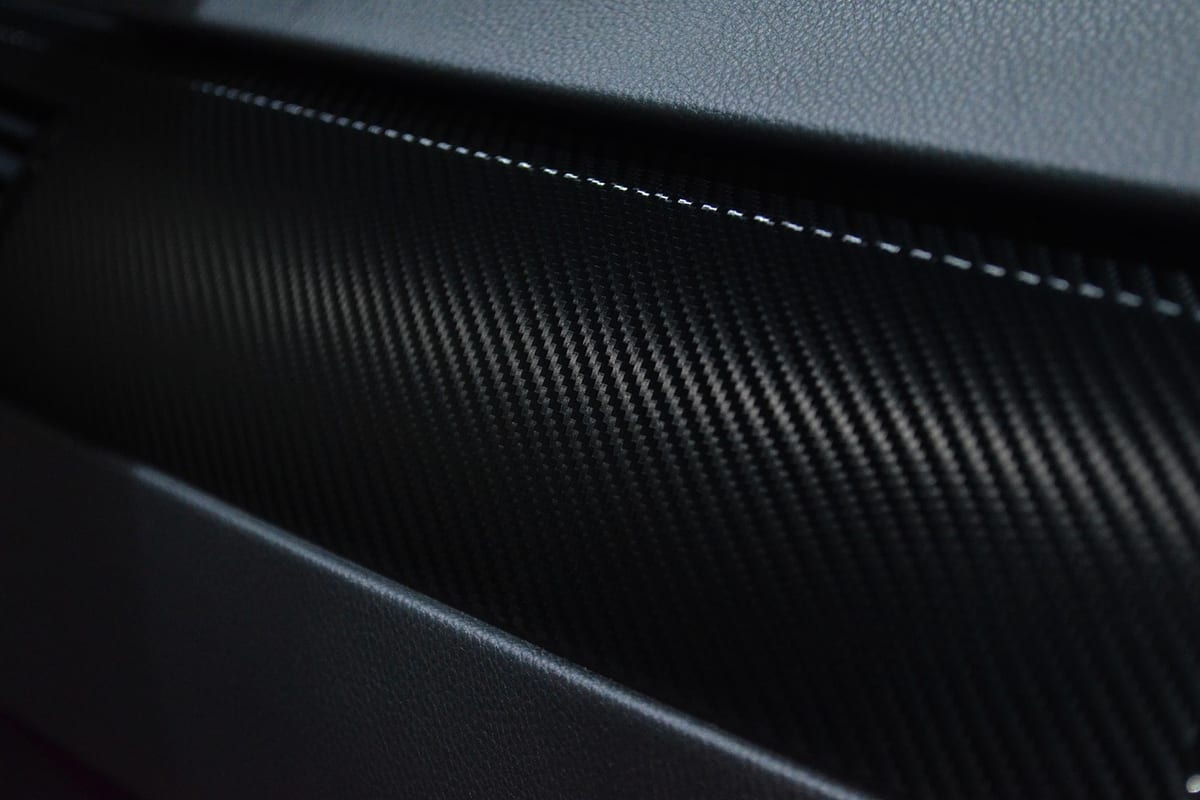 Close-up of black carbon-fiber-pattern vinyl wrap on an interior trim panel, showing the woven texture and a contrast stitch line.