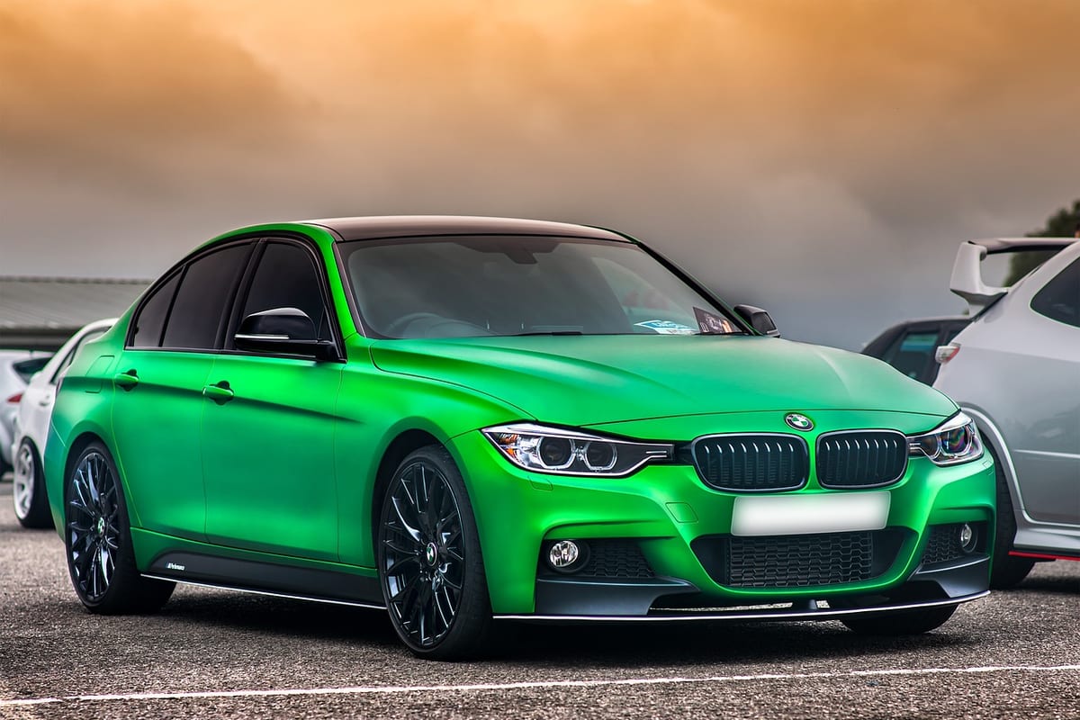 A BMW 3 Series wrapped in a color-shift green vinyl finish, parked outdoors under an overcast sky.